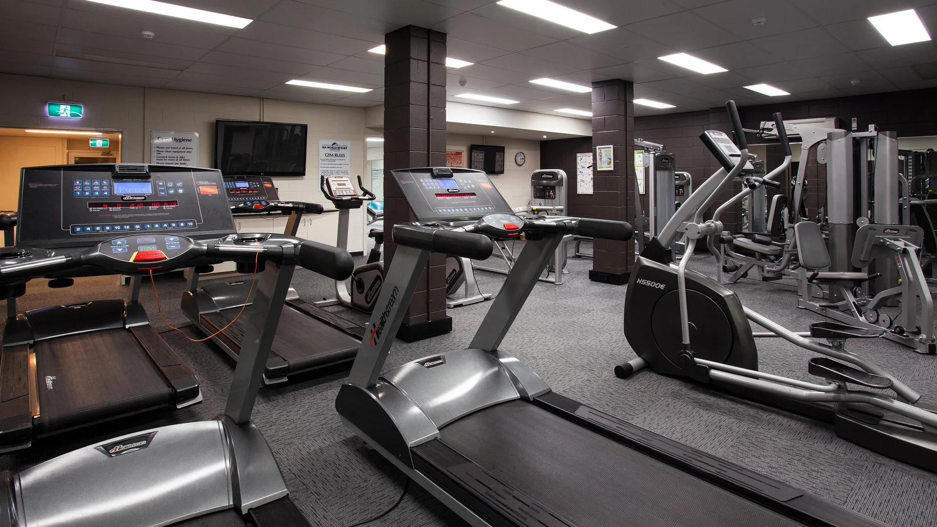 A modern gym with several treadmills, elliptical machines, and various strength-training equipment. The room has grey carpeting, bright lighting, and mirrors on the walls, creating a spacious and clean workout environment.