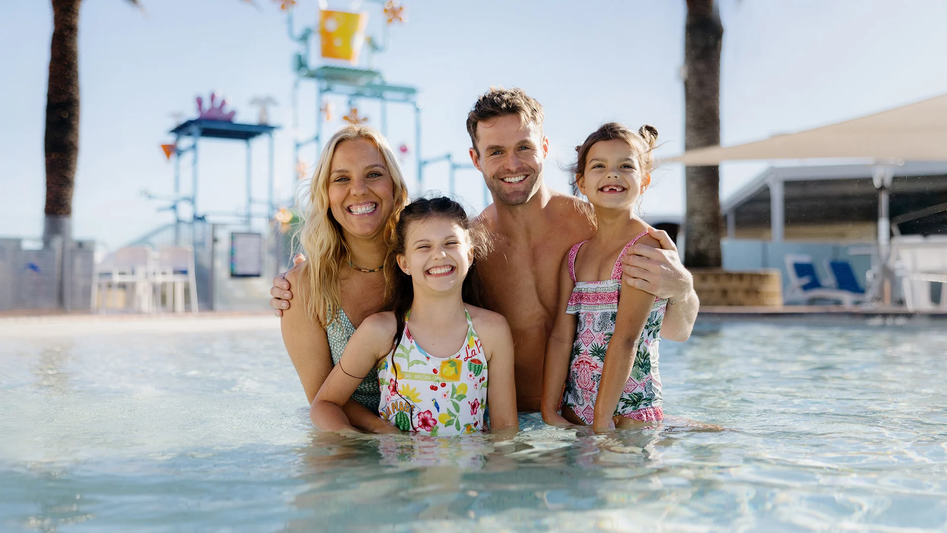A smiling family of four—two adults and two young girls—pose together in a shallow swimming pool at a water park, with colorful water play structures and palm trees in the background.