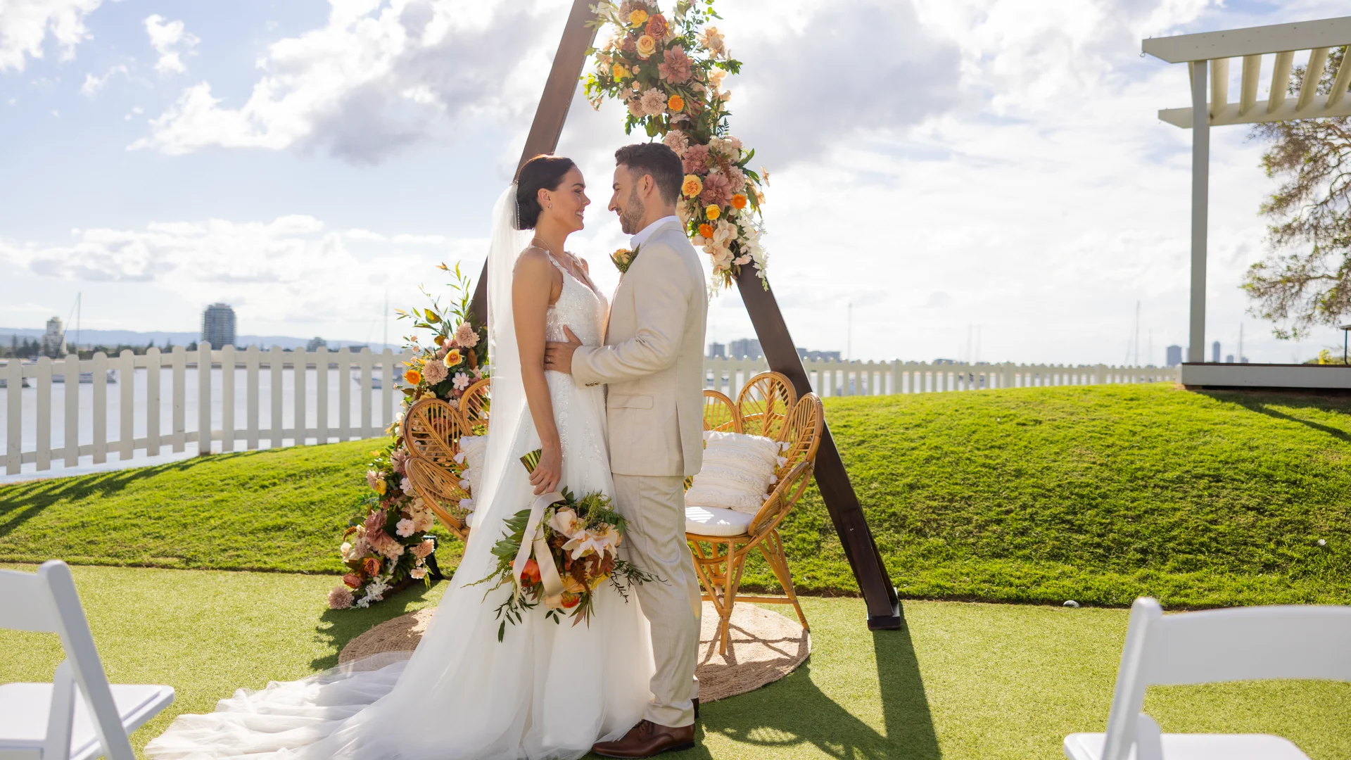 A bride and groom stand facing each other, smiling, in front of a floral wedding arch on a sunny outdoor lawn. White chairs and a city skyline are visible in the background.