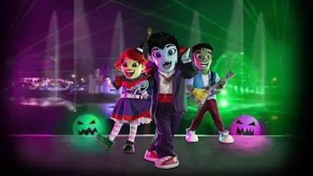 Three colorful, cartoonish monster characters pose on a stage with glowing jack-o'-lanterns and lasers in the background.