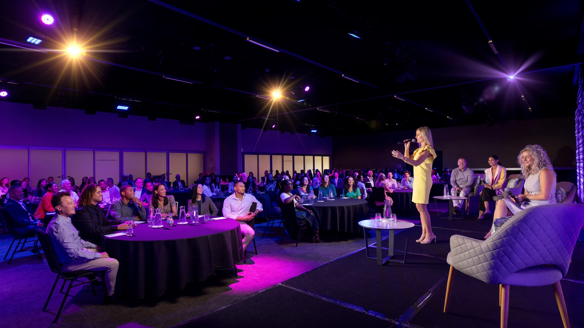 A speaker in a yellow dress addresses an audience in a dimly lit conference room with purple lighting. People are seated at round tables and panel chairs.