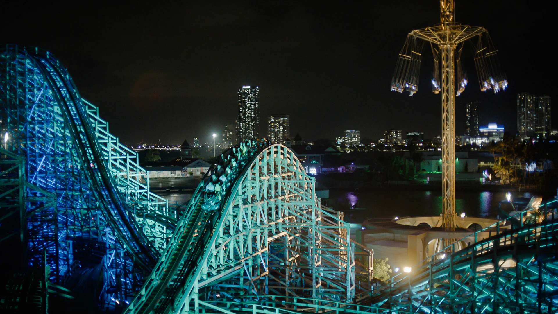 A brightly lit roller coaster and a tall amusement ride are seen at night, with a city skyline and high-rise buildings illuminated in the background.