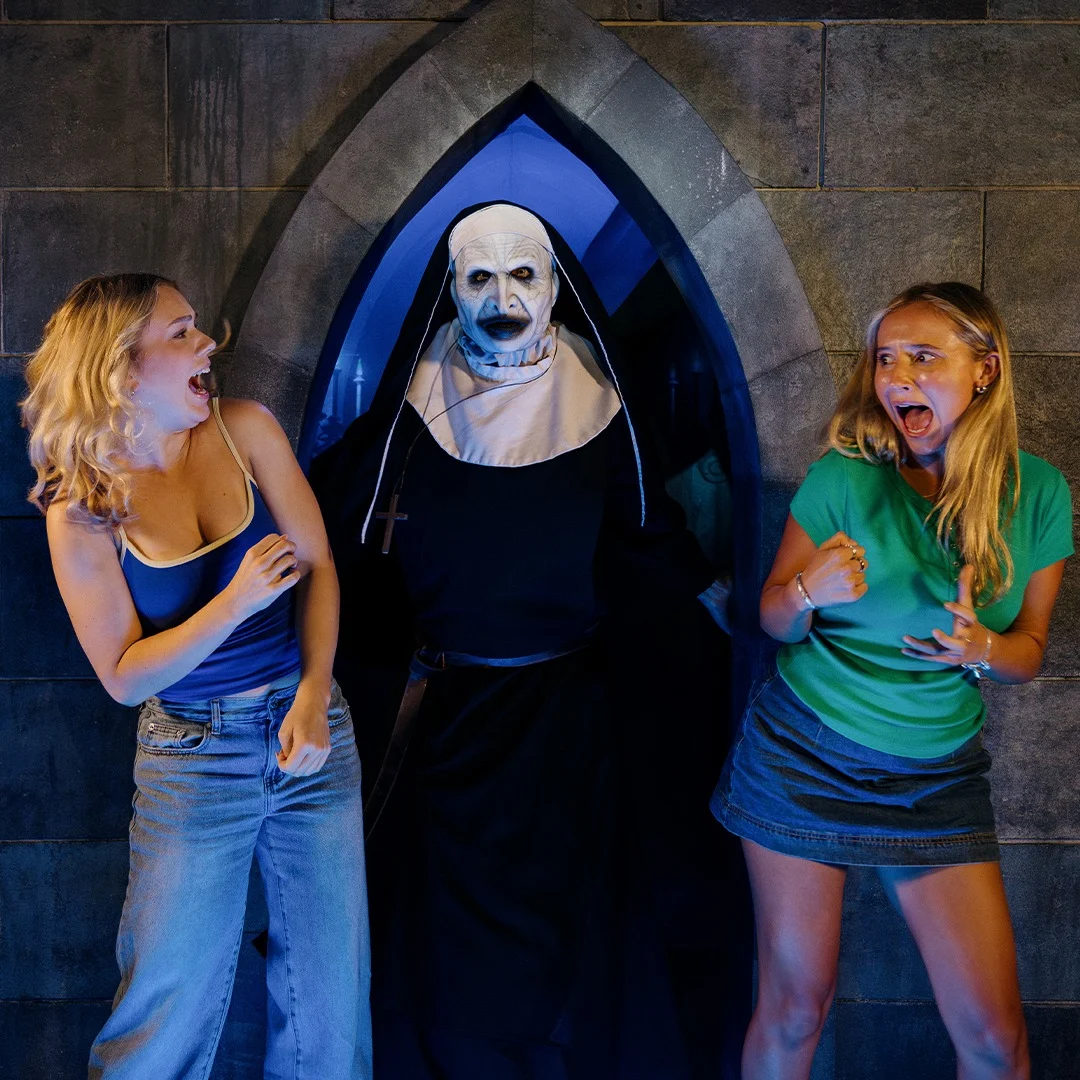 Two women react in fear as a person dressed as a scary nun stands between them against a dark, stone wall with an arched doorway.