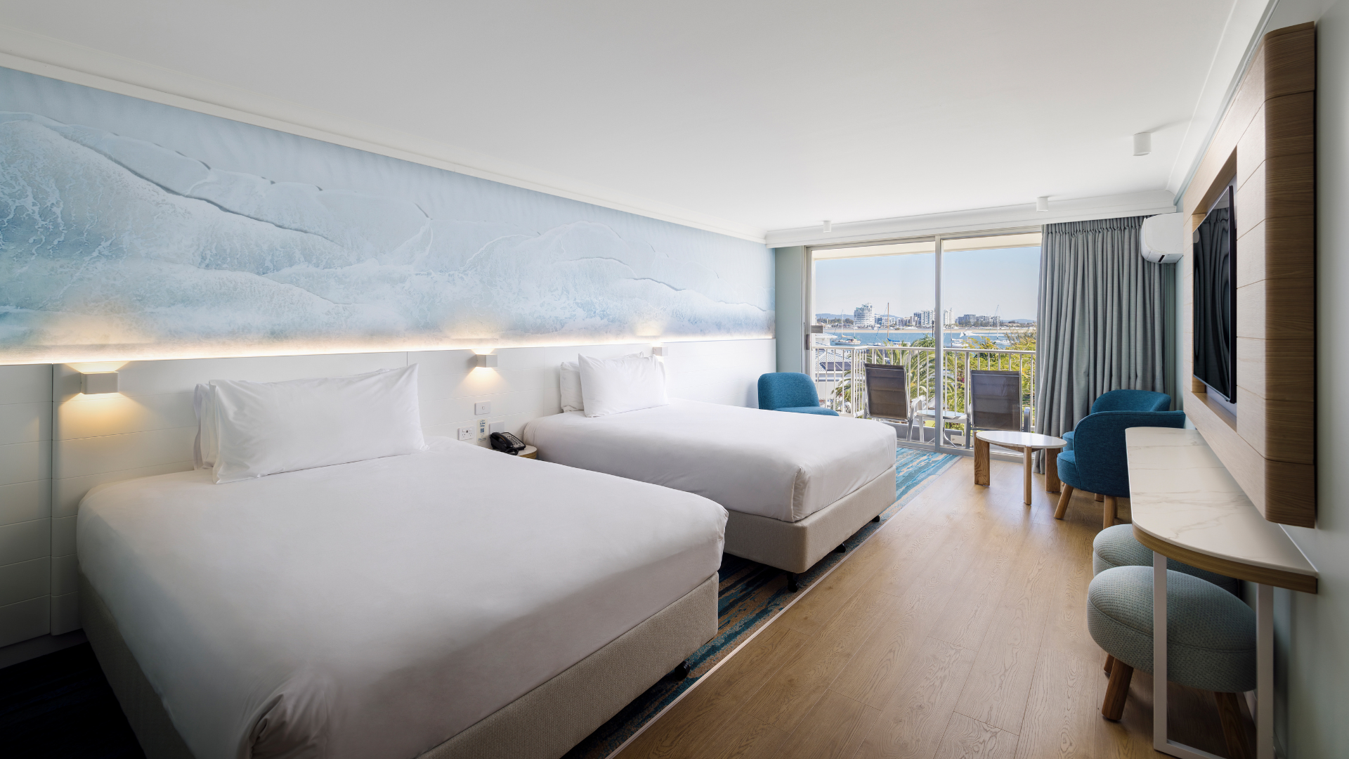 Modern hotel room with two large beds, ocean-themed wall art, a wall-mounted TV, two chairs with a small table, and a balcony overlooking a cityscape.