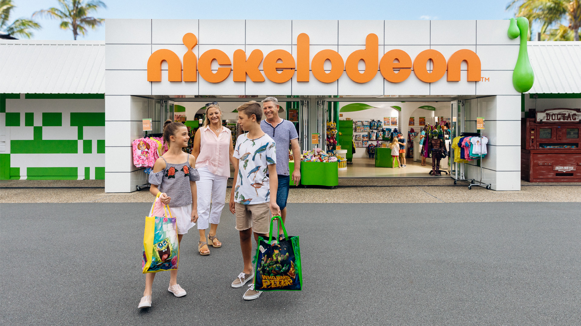 A family of five walks out of a Nickelodeon store, carrying shopping bags and smiling, with merchandise and displays visible inside the store.