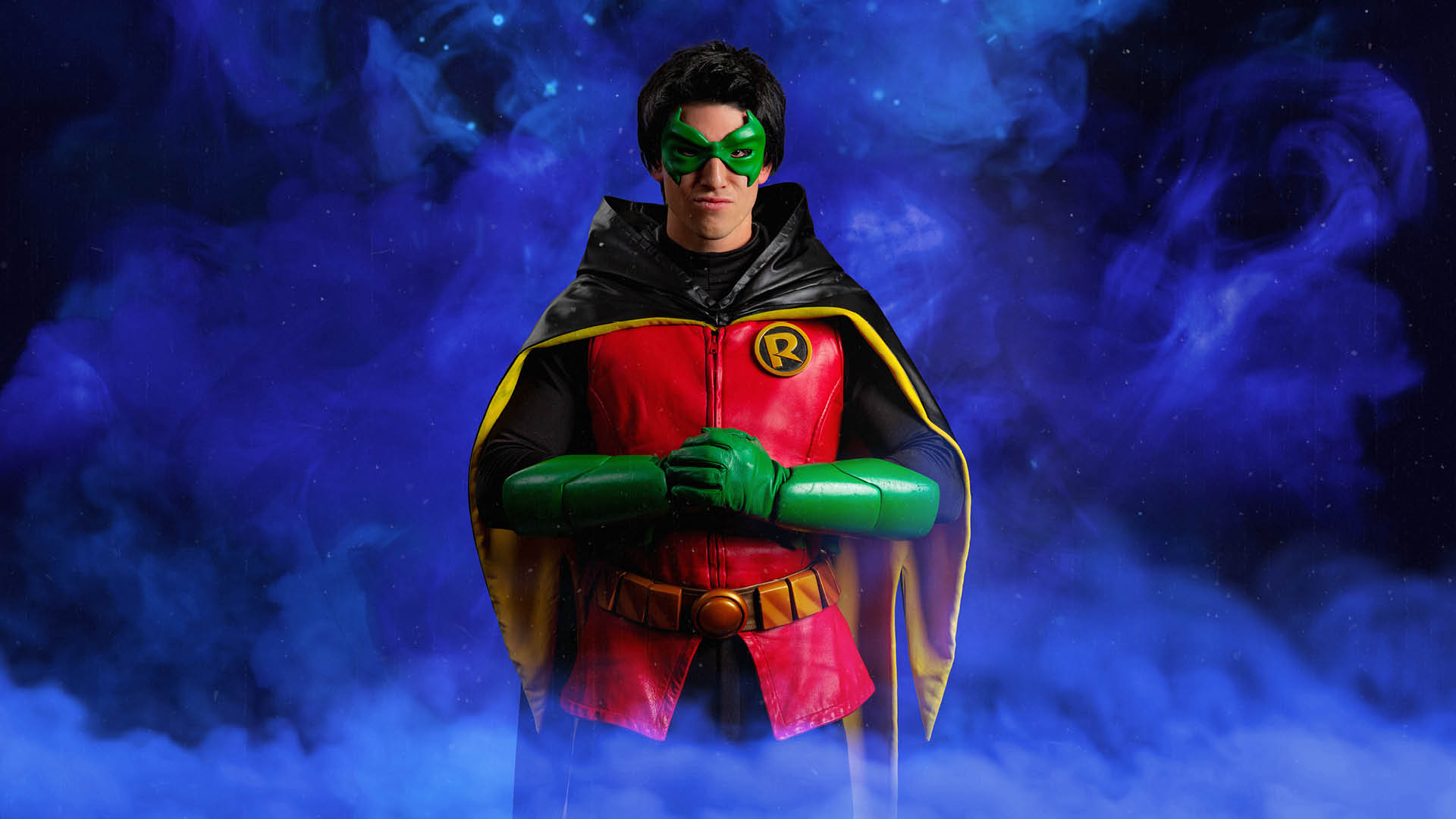 Person dressed as Robin from Batman stands with hands clasped, wearing a red, green, and yellow costume against a purple smoky background.