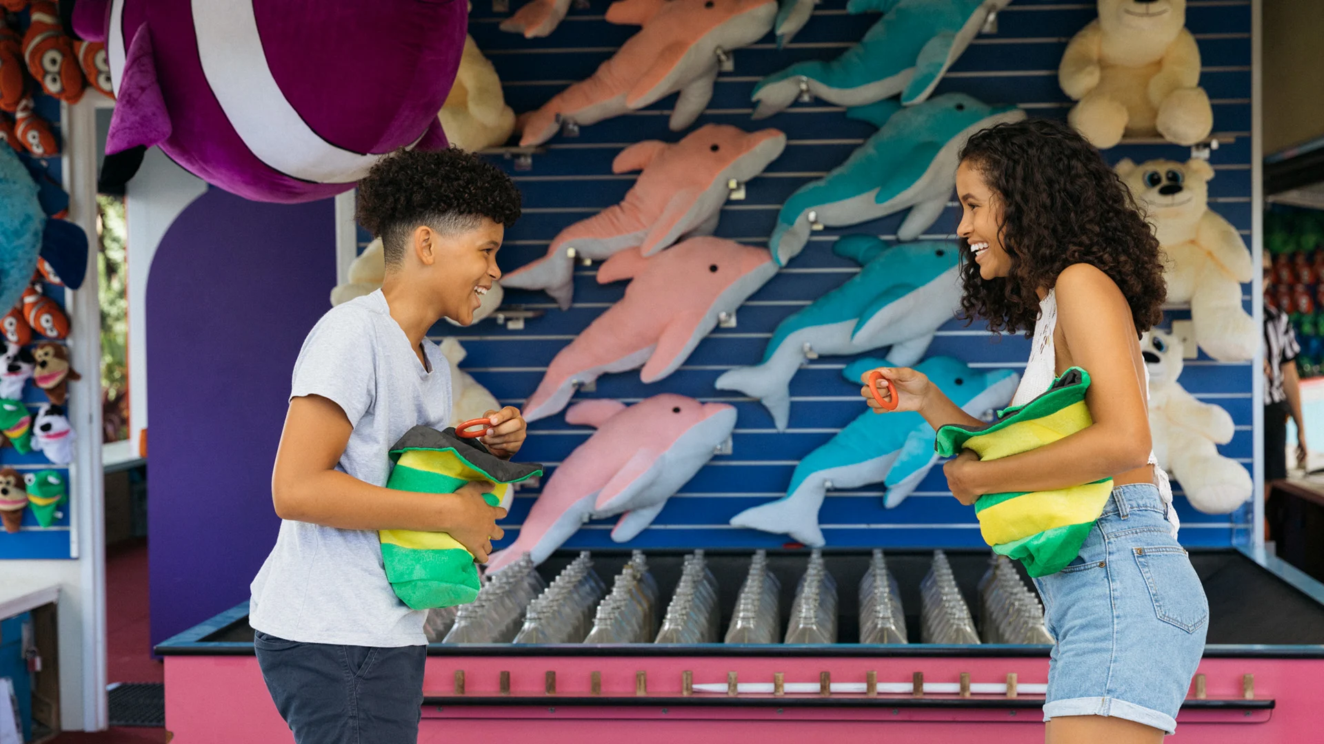 Two people stand at a carnival game booth with stuffed dolphin prizes, each holding a bean bag while smiling at each other.
