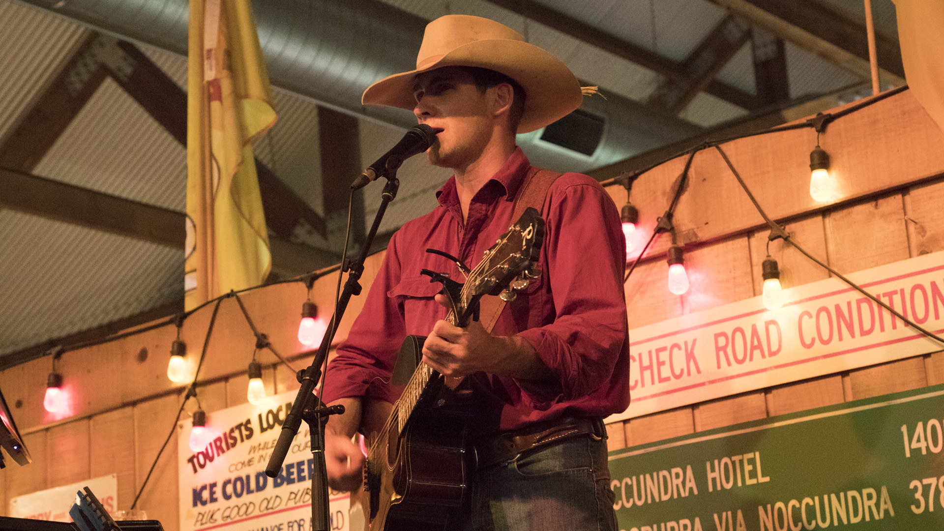 A man in a cowboy hat and red shirt plays an acoustic guitar and sings into a microphone on stage, with colorful string lights and signs in the background.