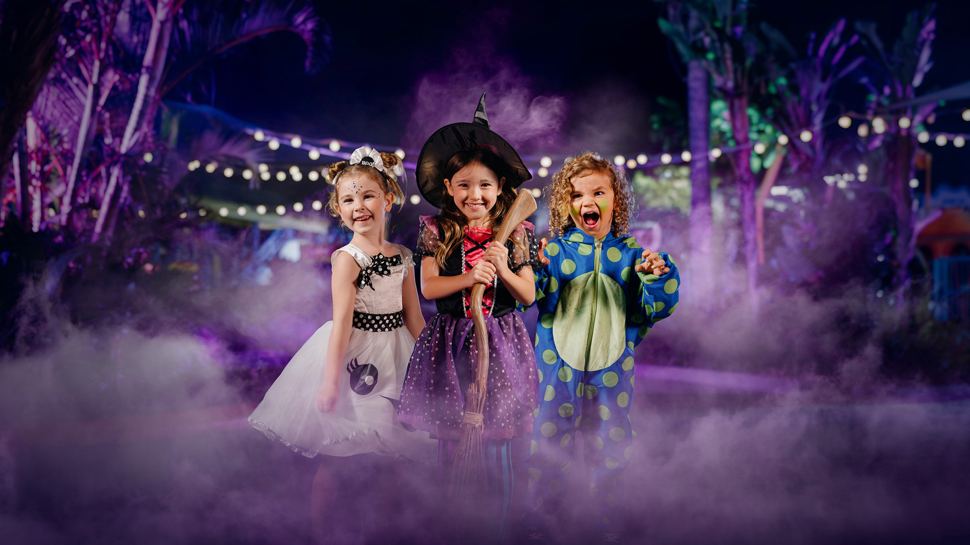 Three young children in Halloween costumes—a ghost, a witch, and a monster—stand together smiling and posing in a misty, outdoor setting with string lights and trees in the background.