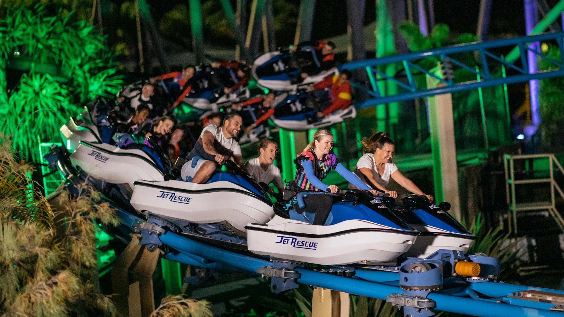 People ride a roller coaster called "Jet Rescue" at night, appearing excited and thrilled as the coaster moves quickly along its blue track surrounded by plants and lights.