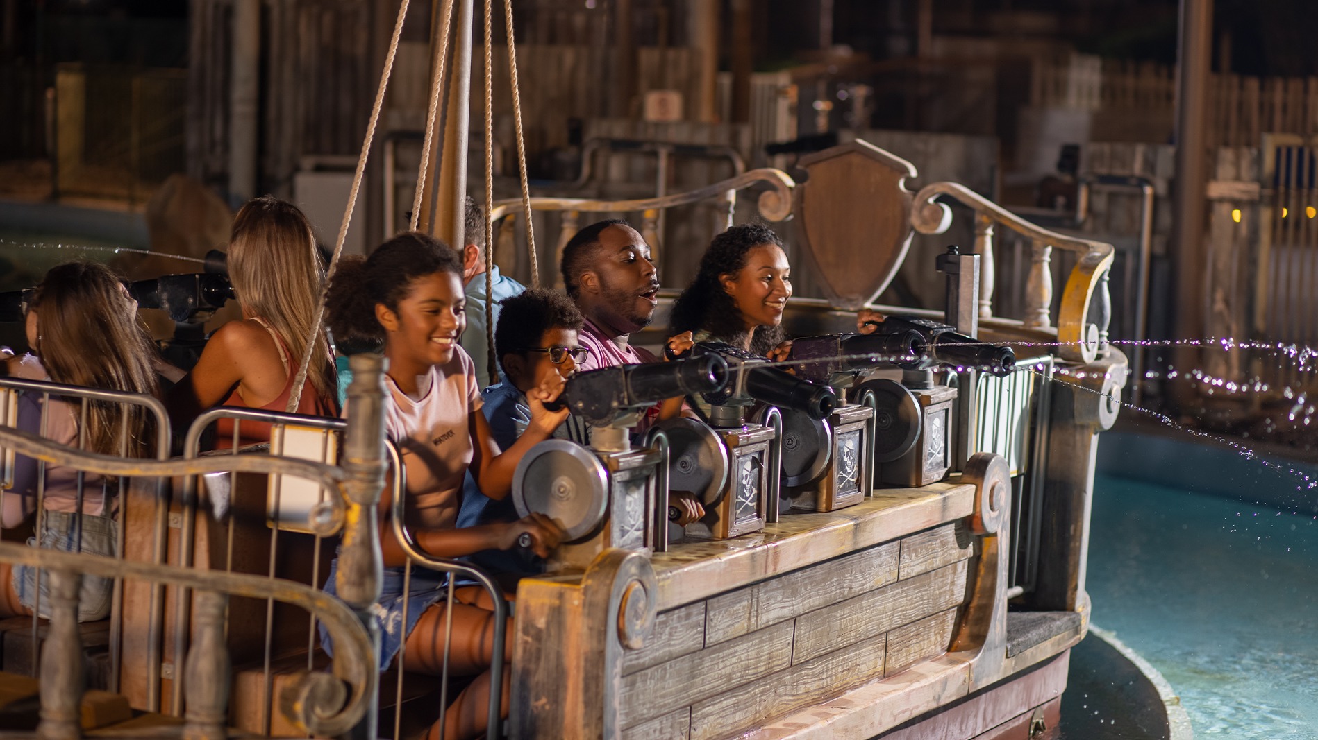 A group of people smiling and laughing while riding a pirate-themed water attraction, aiming water cannons at targets. The scene appears to take place at an amusement park at night.