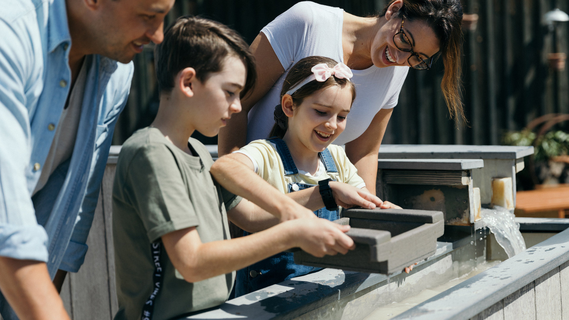 Two children and two adults stand at a water feature, panning for small objects in trays, smiling and engaged in the activity.