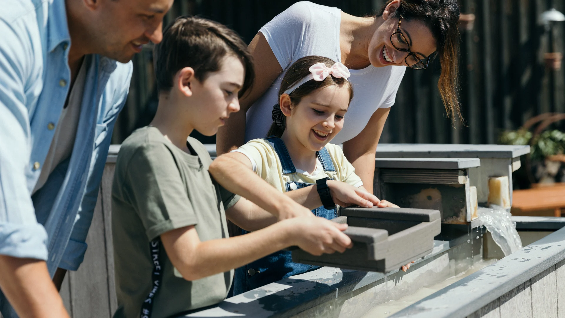 Two children and two adults stand at a water feature, panning for small objects in trays, smiling and engaged in the activity.