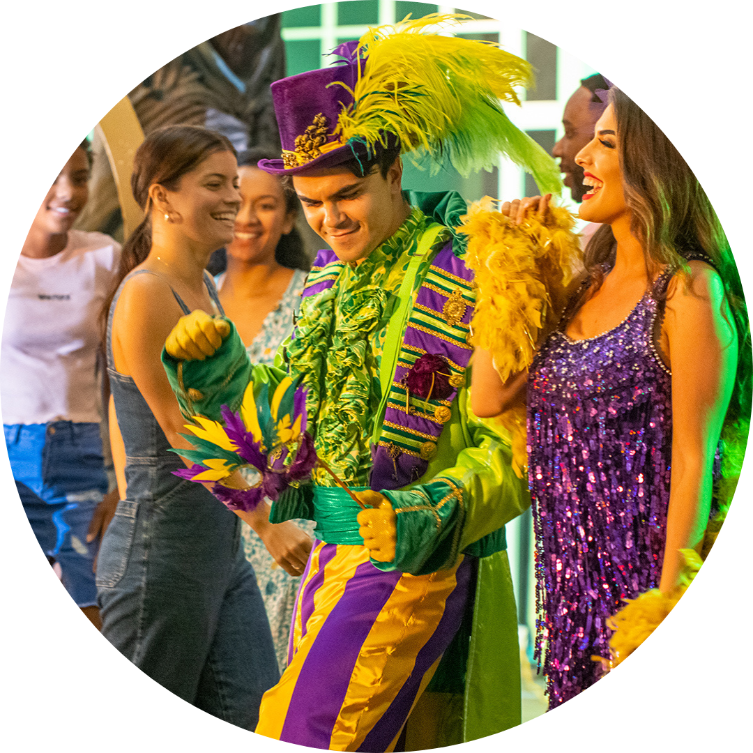 A person in a vibrant Mardi Gras costume with yellow, green, and purple colors and a feathered hat dances with smiling people wearing festive dresses.