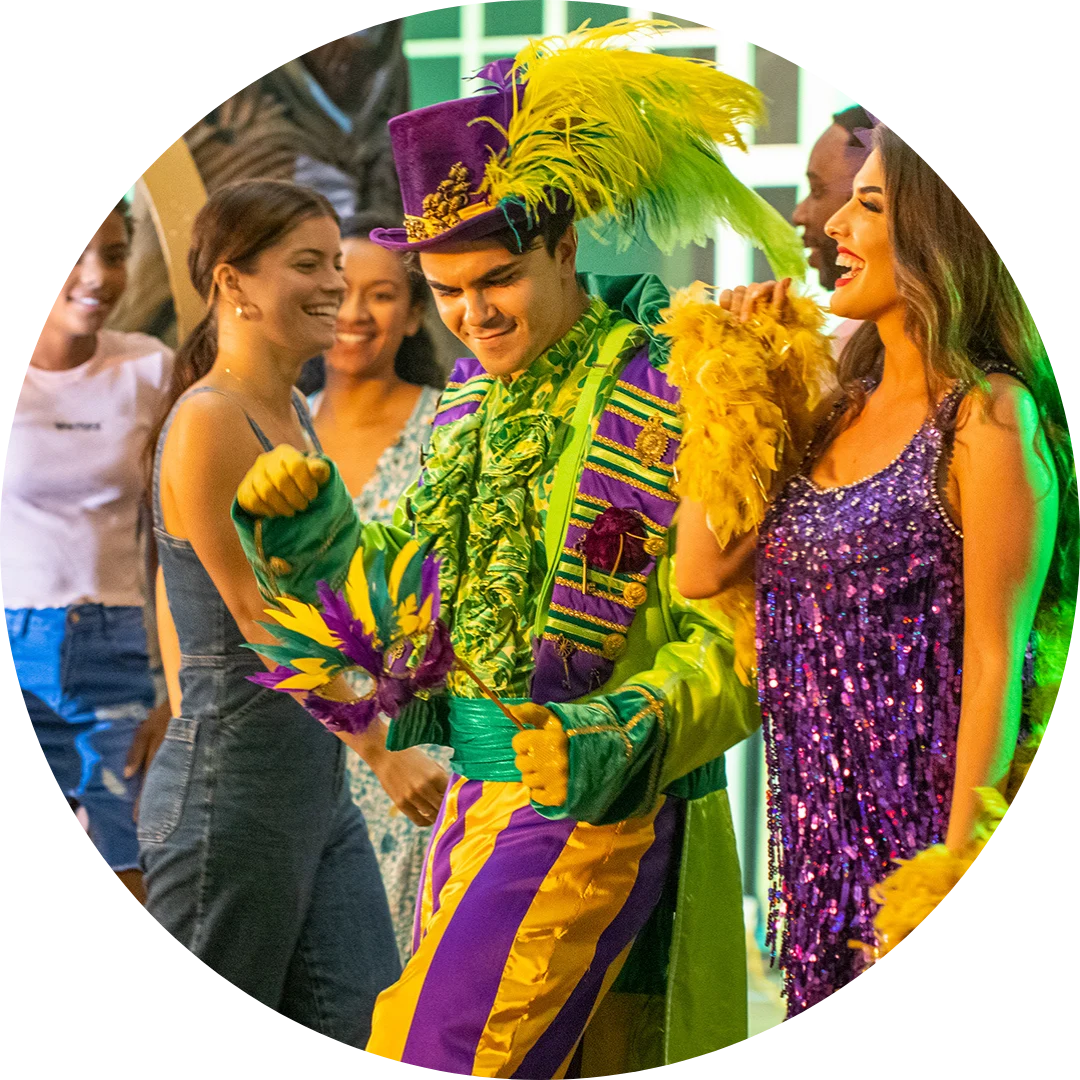A person in a vibrant Mardi Gras costume with yellow, green, and purple colors and a feathered hat dances with smiling people wearing festive dresses.