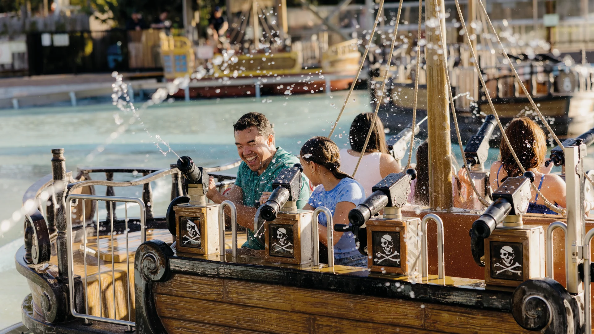 A group of people ride a pirate-themed boat attraction, laughing as water sprays around them. The boat features wooden panels with skull and crossbones decorations. A pool and other rides are visible in the background.