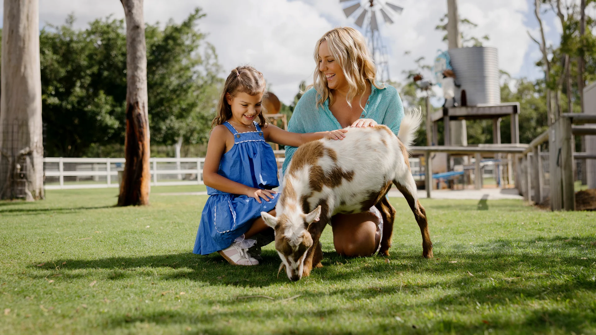 A smiling woman and young girl kneel on grass, petting a small brown-and-white goat. The goat grazes as they watch. A windmill, wooden structures, and trees are visible in the background under a partly cloudy sky.
