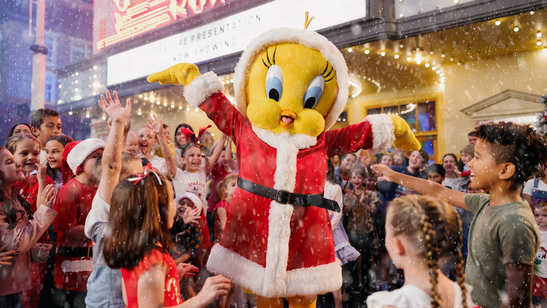 Tweety Bird, dressed in a red Santa outfit, stands with arms outstretched, surrounded by smiling children in festive attire, as artificial snow falls in a holiday-themed outdoor setting.