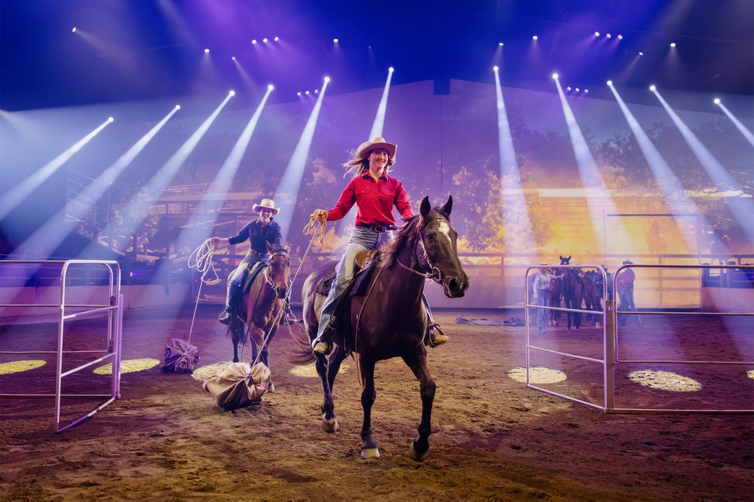 Two cowboys on horseback perform under bright stage lights in an indoor arena, with metal gates and other performers visible in the background.