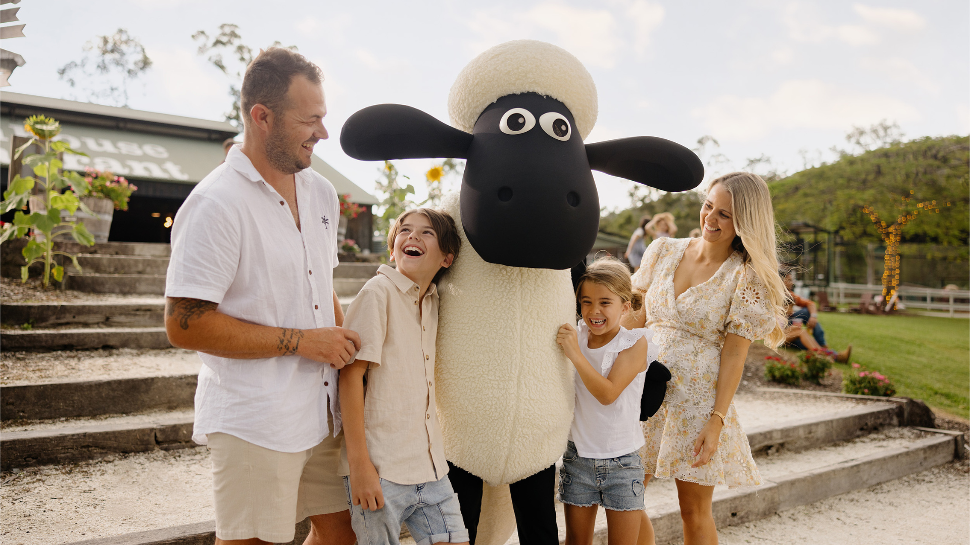 A smiling family of four poses outdoors with a person in a large Shaun the Sheep costume. The parents and two children stand close together, laughing and enjoying a sunny day.