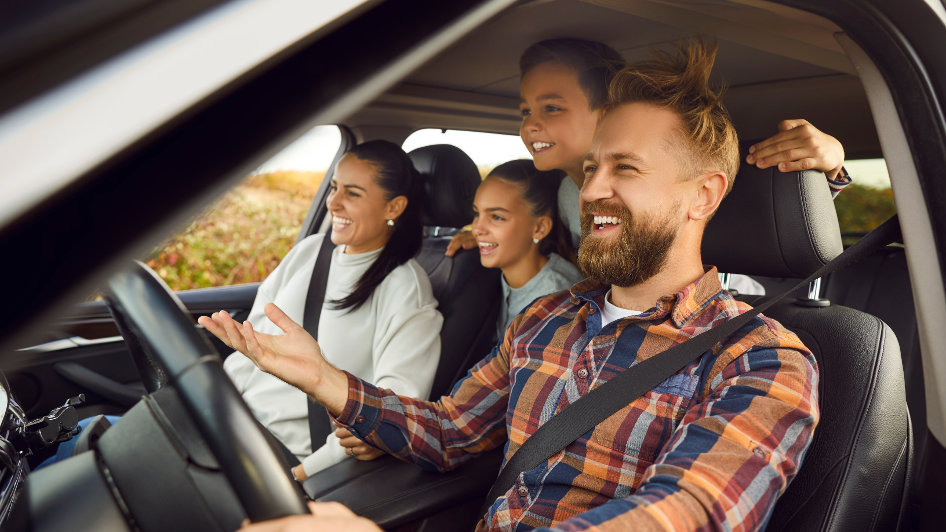 A smiling family of four is inside a car, with the father driving and others sitting beside and behind him.