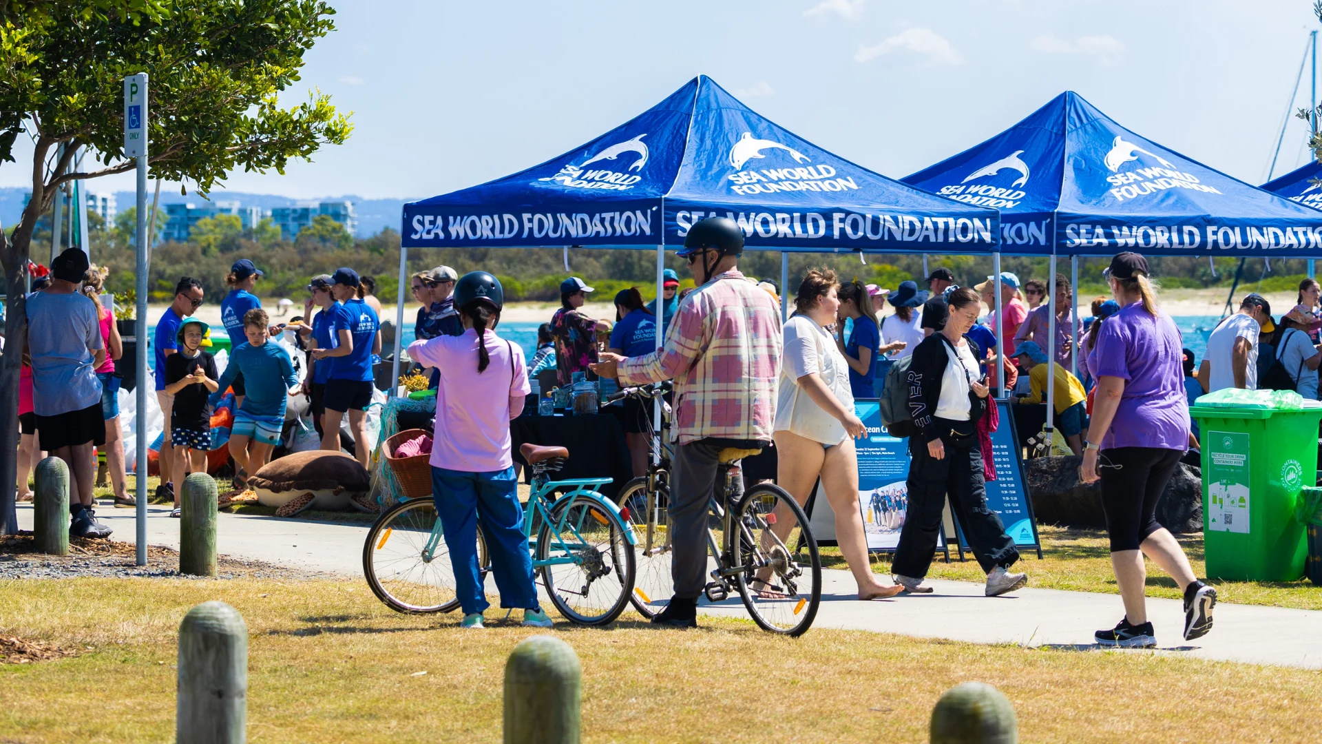A crowd gathers at an outdoor event near blue Sea World Foundation tents. People walk, ride bikes, and talk, while volunteers assist near tables. It’s a sunny day with trees and blue sky in the background.