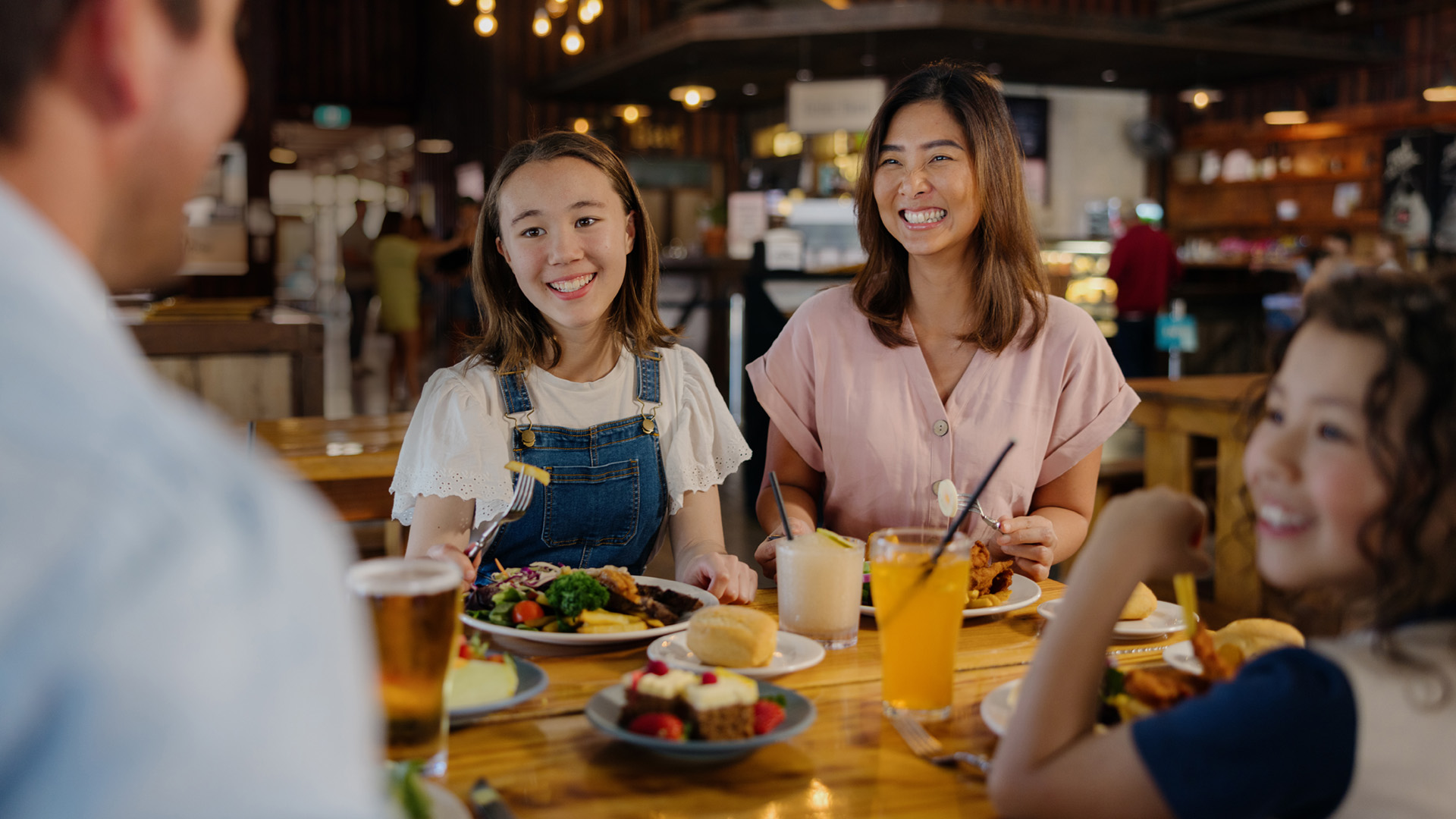 Four people sit at a wooden table in a restaurant, smiling and enjoying a meal with drinks, salad, fruit, and dessert.