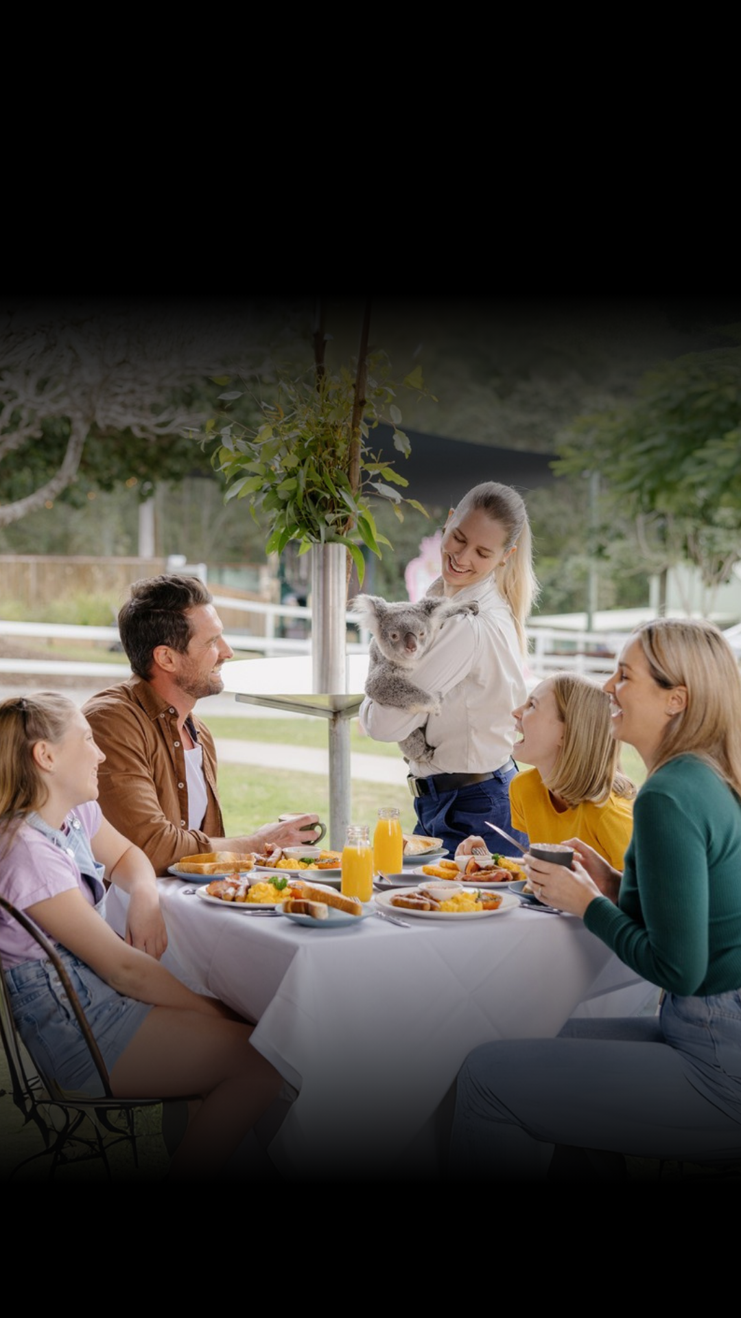 A family of five sits at an outdoor table with breakfast food, while a girl stands holding a koala.
