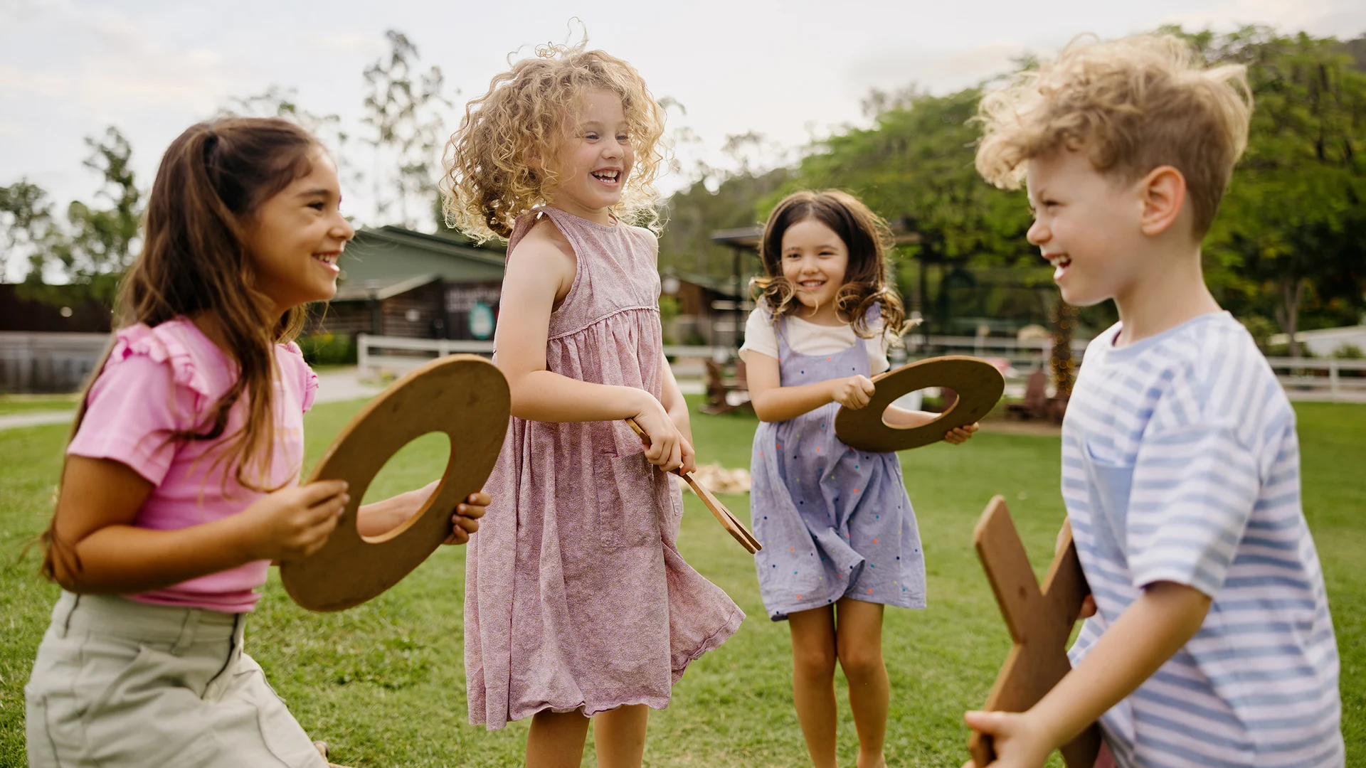 Four young children play and laugh together outdoors on grass, holding round wooden pieces. They appear happy and energetic, with trees and buildings visible in the background on a sunny day.