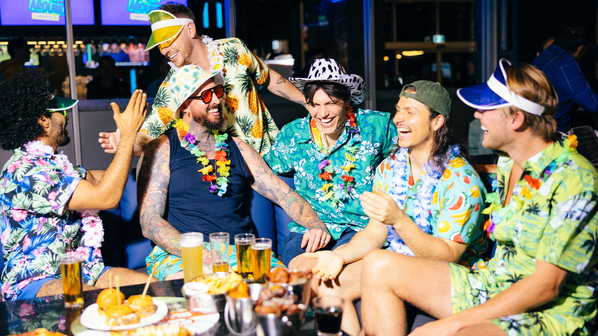 Six men wearing colorful tropical shirts, leis, and visors are laughing and high-fiving around a table with drinks and snacks in a lively, brightly lit indoor setting.