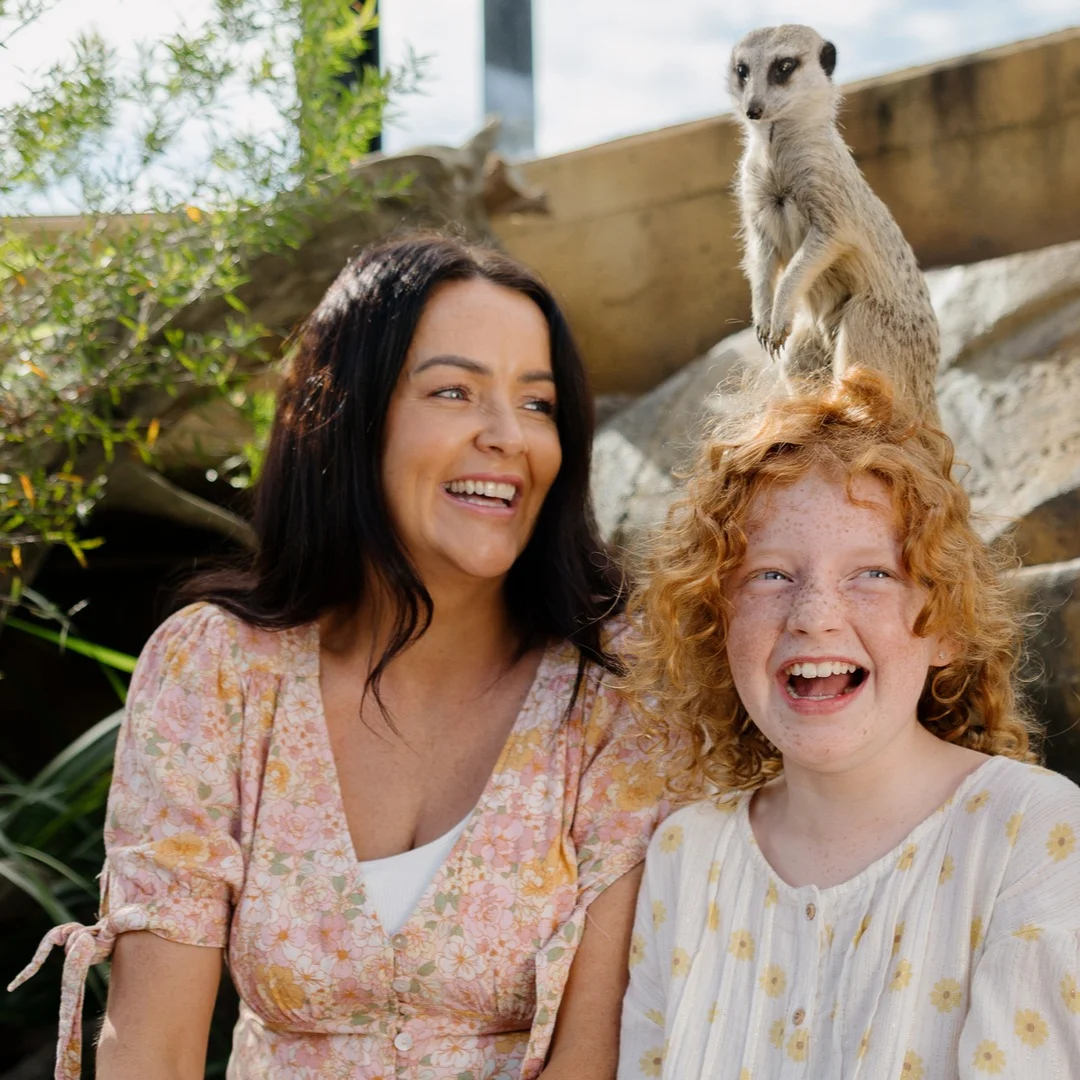 Two people, a woman and a child with curly red hair, are smiling and laughing outdoors. A meerkat is perched on a rock directly behind them, appearing to sit on the child’s head. The scene is bright and cheerful.