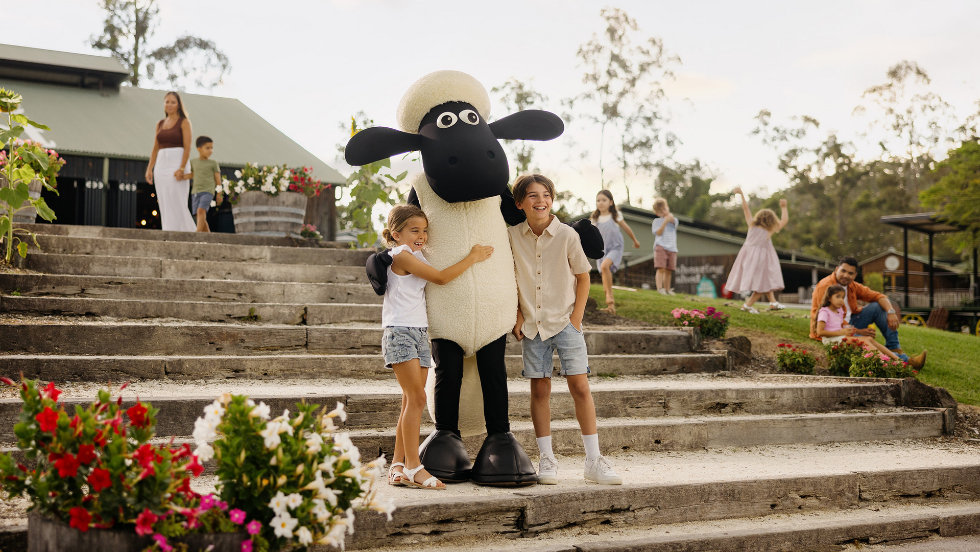 Two children hug a person in a large Shaun the Sheep costume on stone steps surrounded by flowers, while families and children gather and relax in the background at a park or outdoor event.