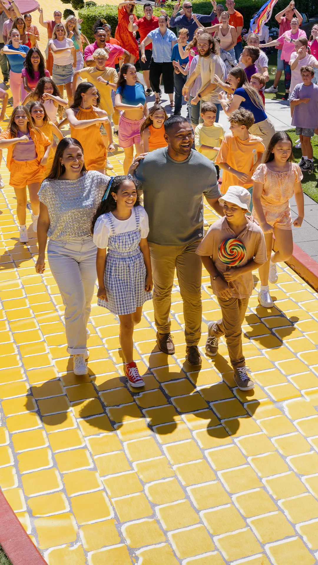 A group of people, including a family of four, walk together on a yellow brick path outdoors. The family is at the center, smiling and holding hands, with children and adults around them in colorful clothing.