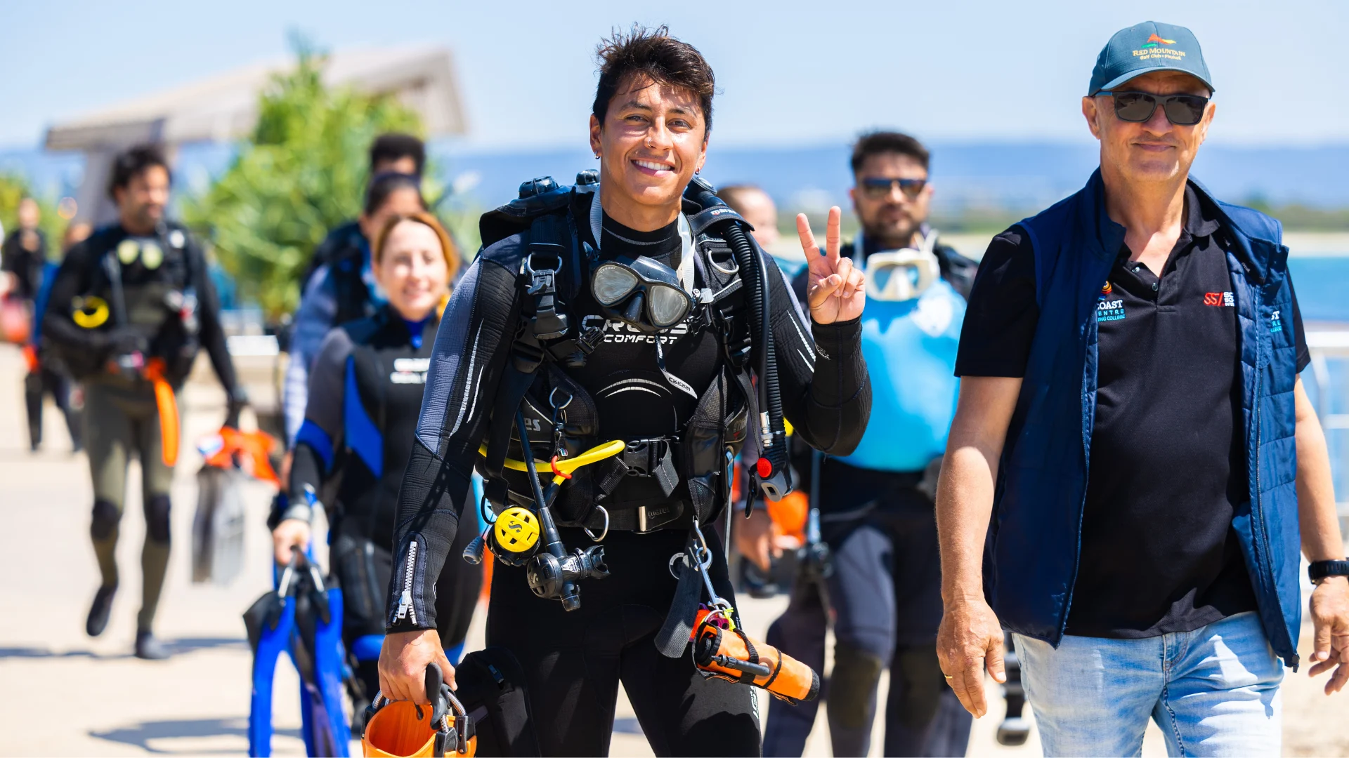 A group of scuba divers in wetsuits and gear walk along a sunny waterfront path, with a smiling man at the front flashing a peace sign. A man in a cap and sunglasses walks beside them.