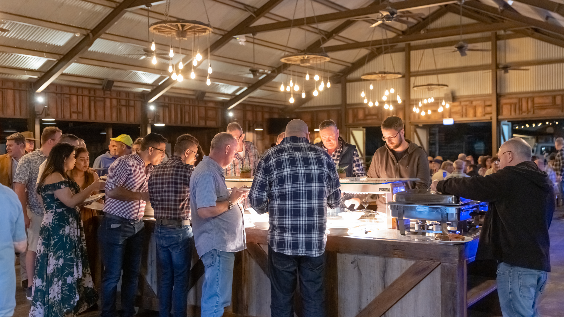 A group of people serve themselves food from a buffet inside a rustic, well-lit event hall with wooden beams and hanging lights.