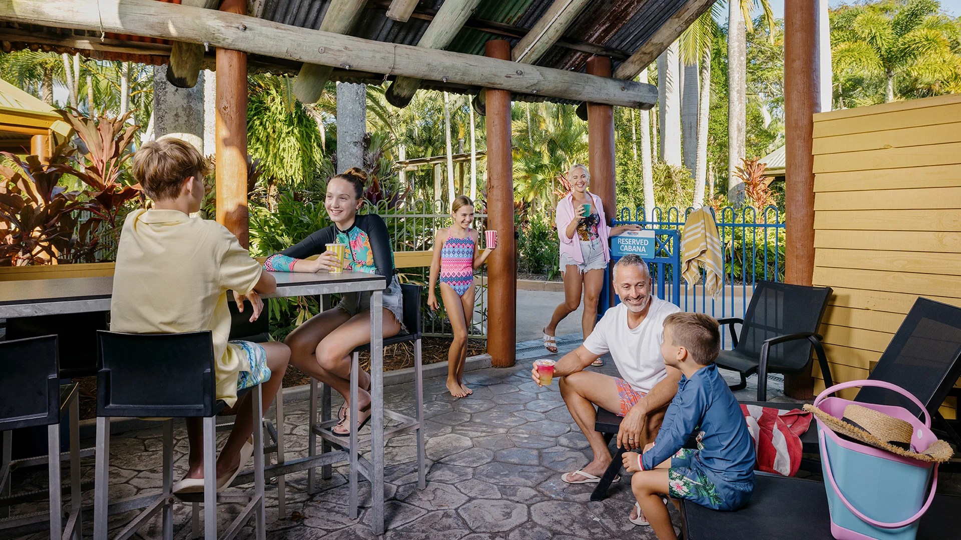 Group of people, including children, relaxing at an outdoor bar area near a pool. Some are seated, holding drinks, while others stand. Palm trees and tropical plants are in the background.
