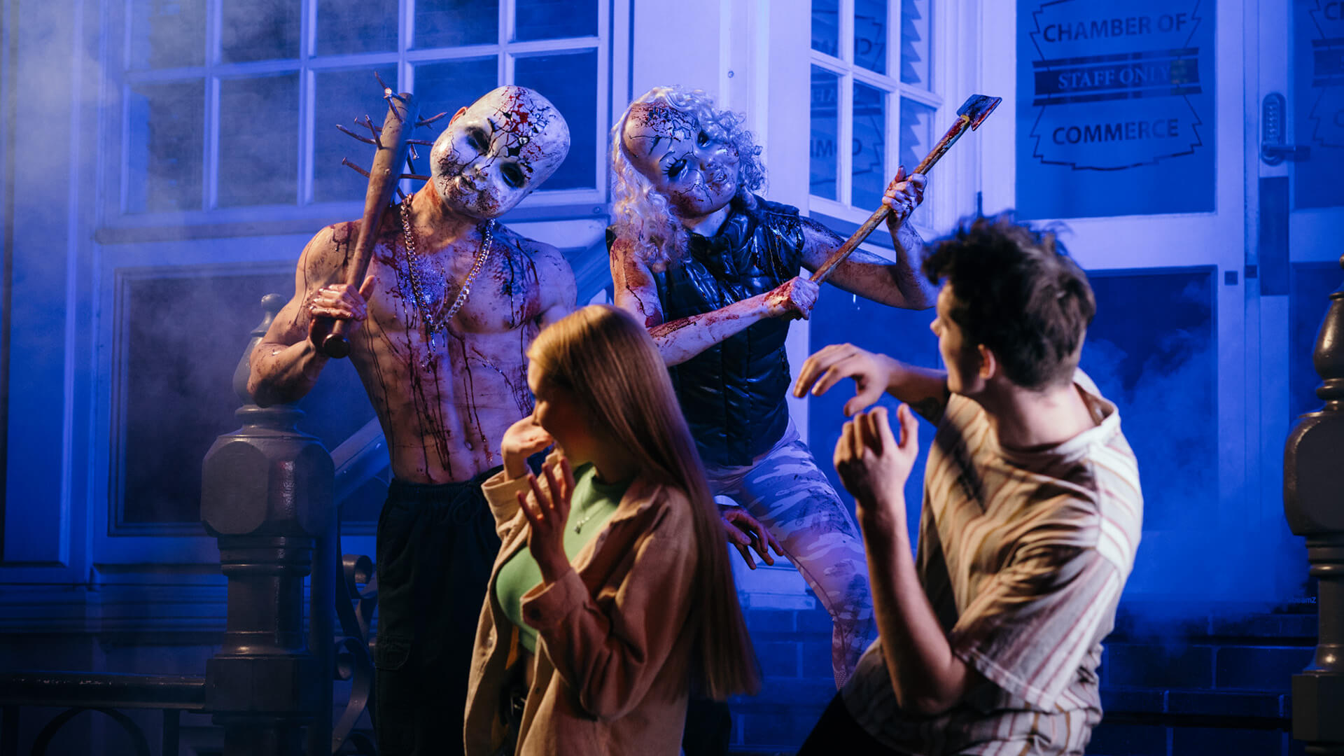 Two people in costumes and scary masks holding bloody weapons approach a frightened man and woman outside a building at night, creating a horror-themed scene with dramatic lighting and fog.