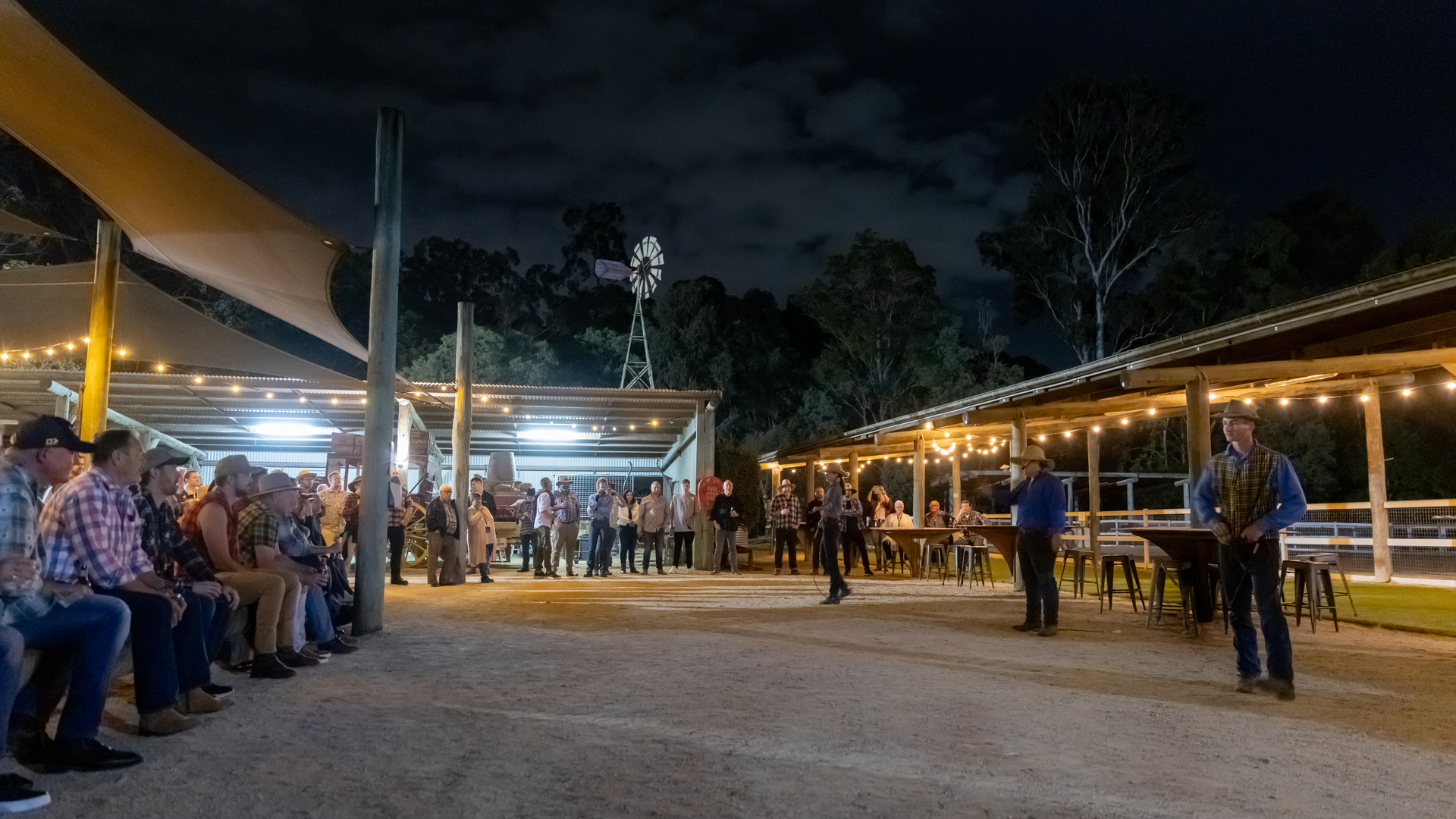 A group of people sits and stands under string lights at an outdoor venue at night, with trees and a windmill visible in the background.