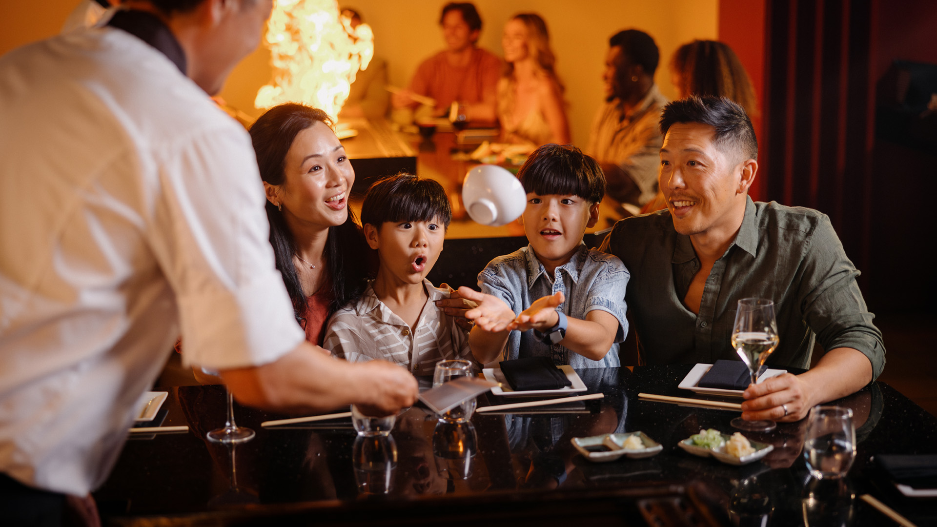 A family of four sits at a teppanyaki restaurant table, reacting with excitement as a chef performs a cooking trick with flames in the background. The children look amazed, and the parents smile while holding drinks.