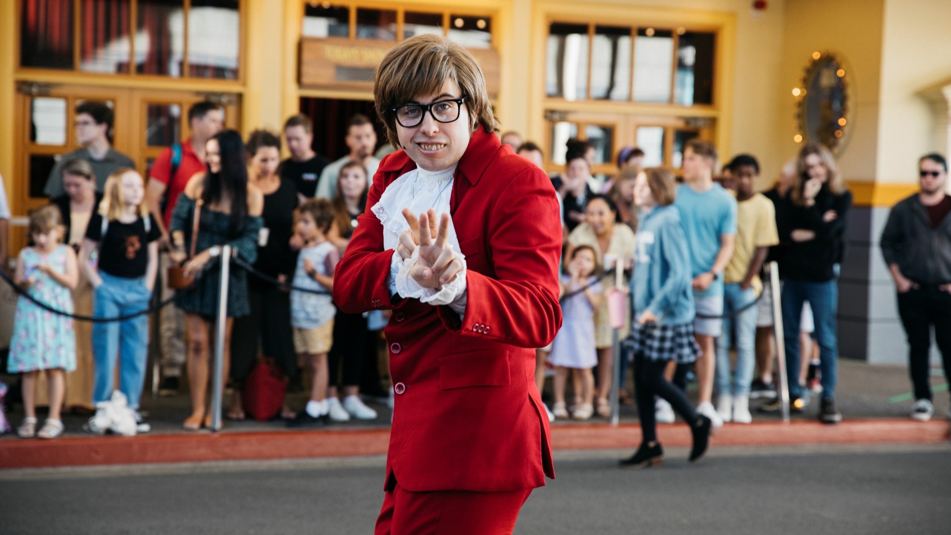 Person dressed as Austin Powers in a red suit poses with a peace sign gesture outside, while a crowd of people stands in the background.