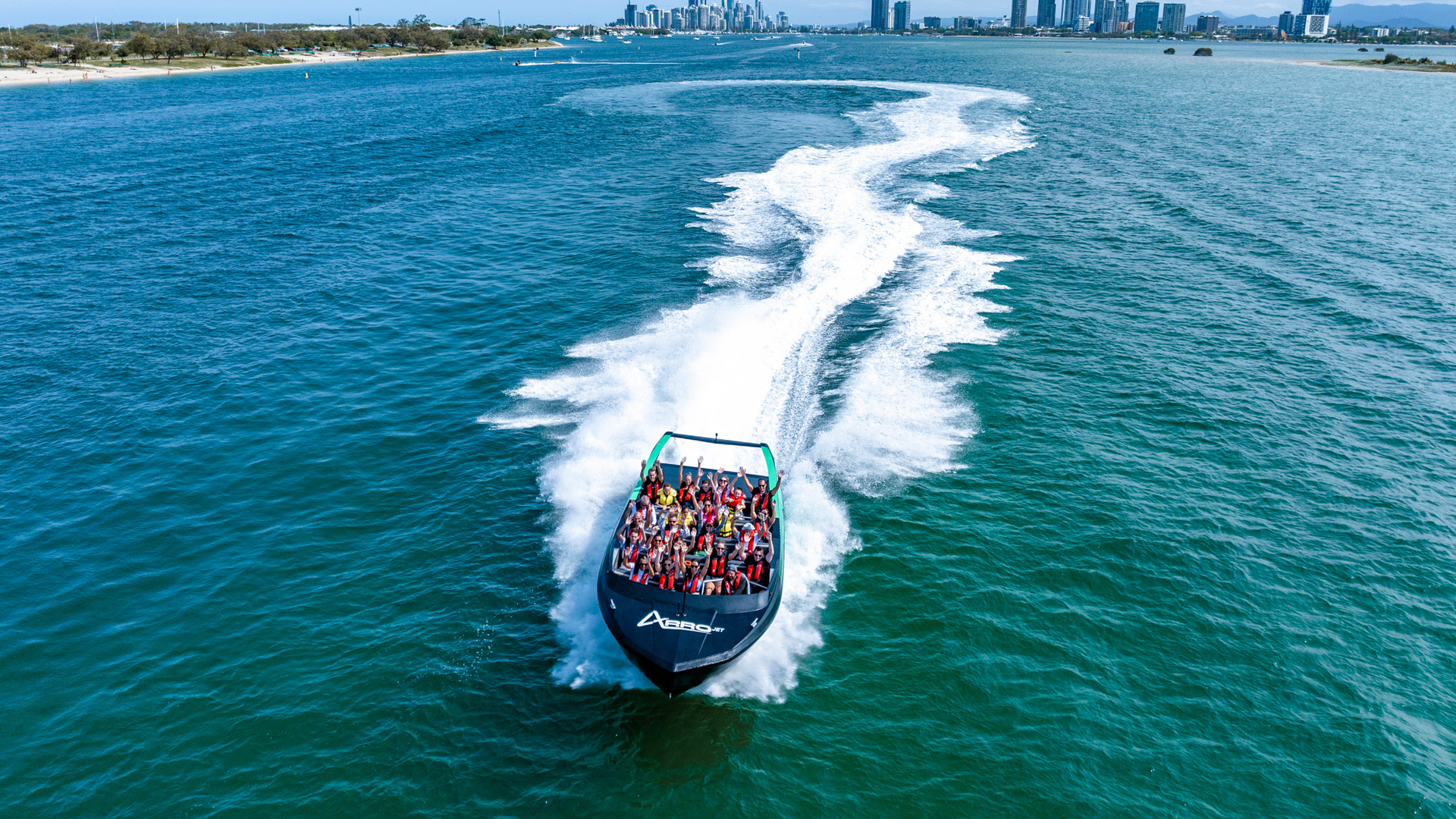 A speedboat filled with people creates a white wake as it travels quickly across blue water, with a city skyline and tall buildings visible in the distant background.