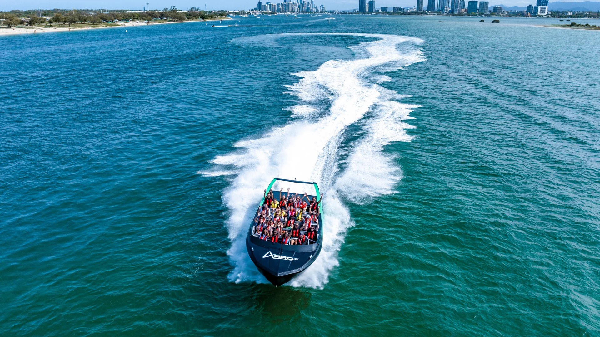 A speedboat filled with people creates a white wake as it travels quickly across blue water, with a city skyline and tall buildings visible in the distant background.