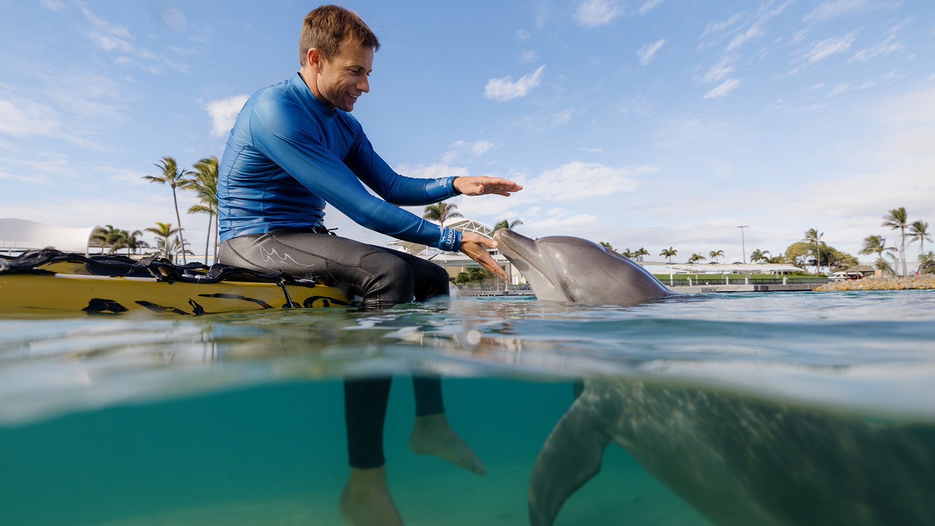 Man in blue shirt sitting on a kayak interacting with a dolphin in clear water, under a partly cloudy sky.