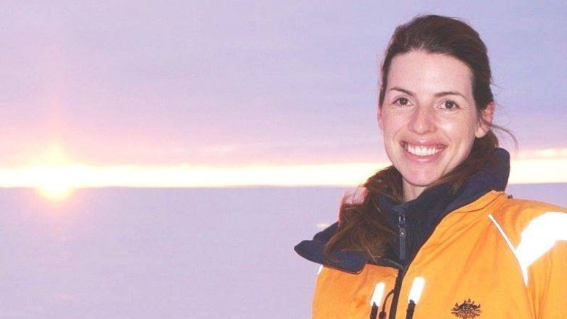 A woman in a yellow jacket stands outdoors in front of a snowy landscape with the sun low on the horizon.