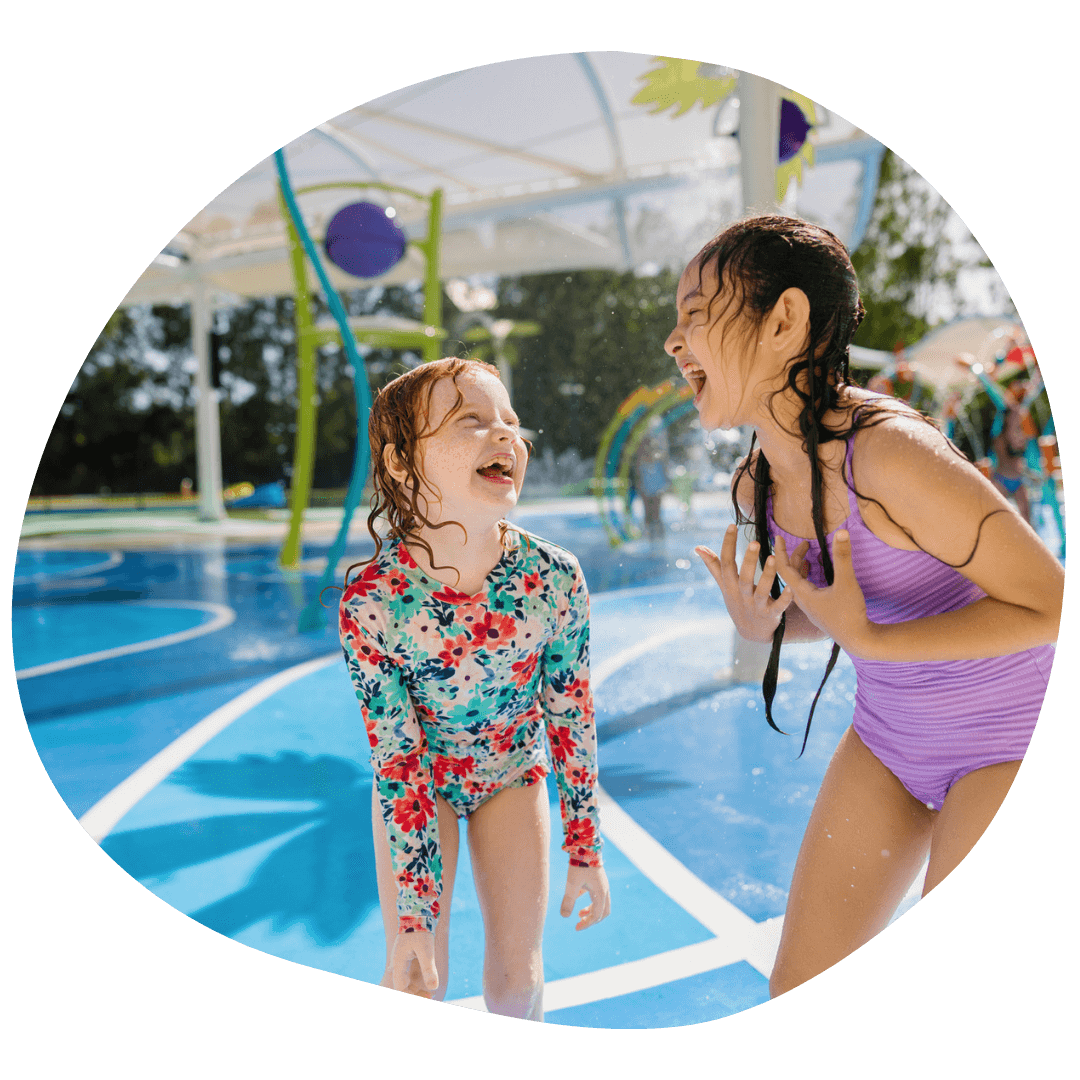 Two children laughing and playing at a water park, standing on a wet surface under a sunny sky, with water sprays in the background.