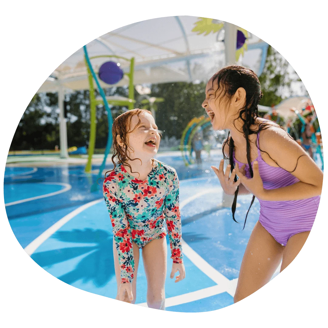Two children laughing and playing at a water park, standing on a wet surface under a sunny sky, with water sprays in the background.