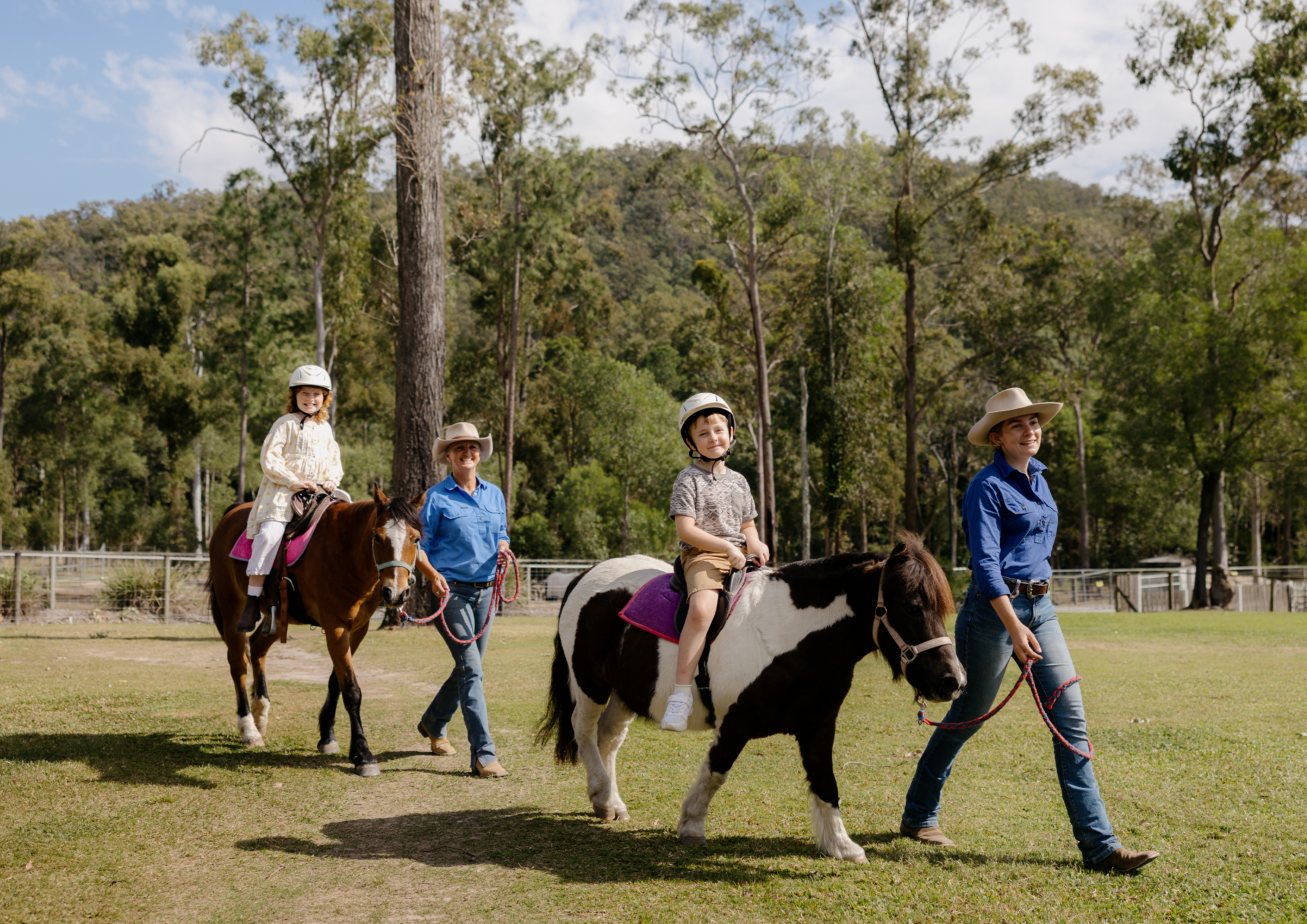 Two adults lead two children on horseback through a grassy area with trees and hills in the background. The children wear helmets, and everyone appears to be enjoying a sunny day outdoors.