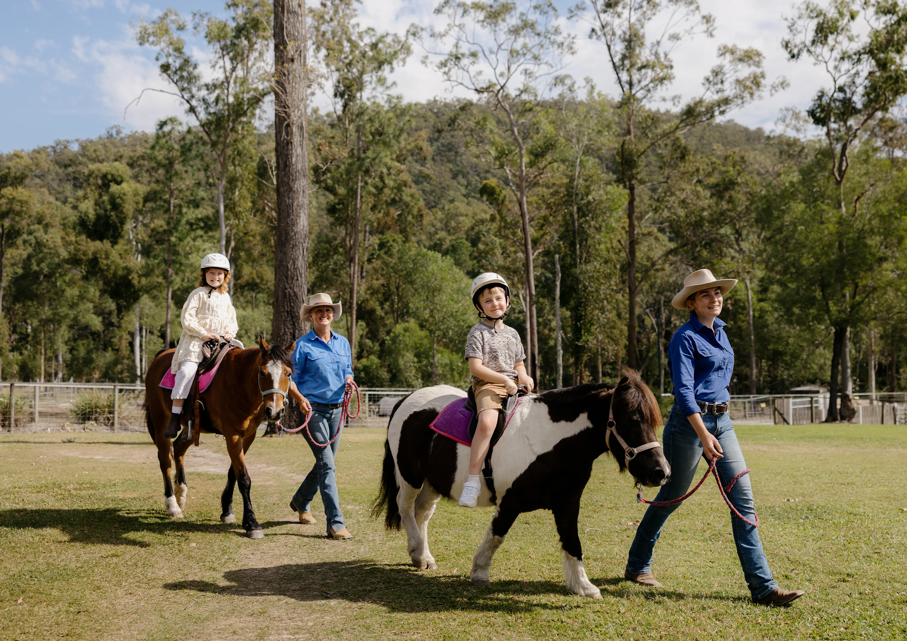 Two adults lead two children on horseback through a grassy area with trees and hills in the background. The children wear helmets, and everyone appears to be enjoying a sunny day outdoors.