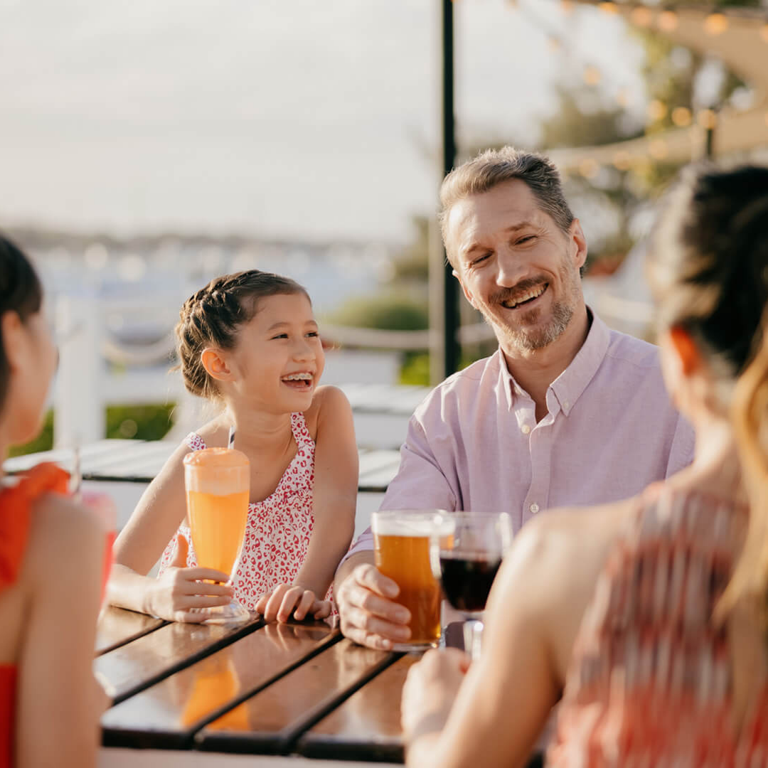 A smiling family sits at an outdoor table, enjoying drinks together. A young girl and an adult man face the camera, both laughing, while two other people are seen from behind. Trees and water are visible in the background.