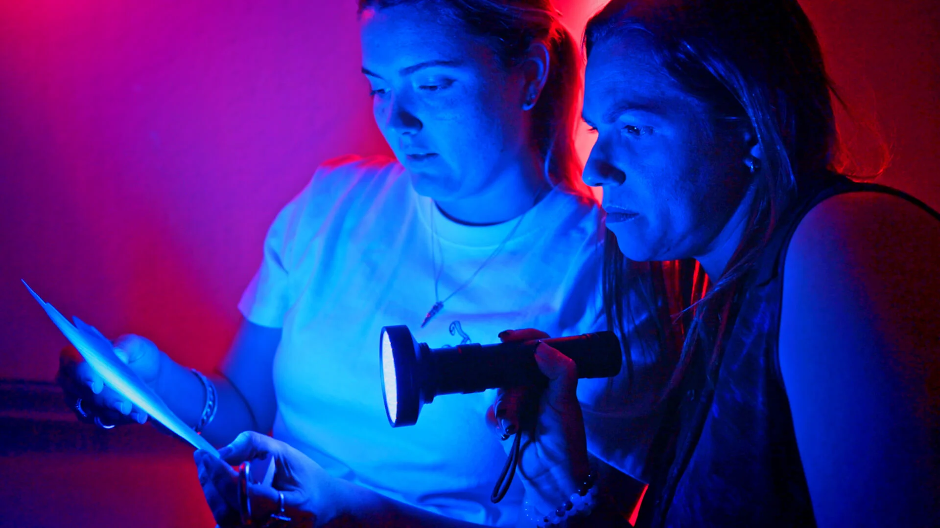 Two people reading a piece of paper solving clues under glowing red and blue lights with a torch inside the Warner Bros. Movie World escape room attraction.